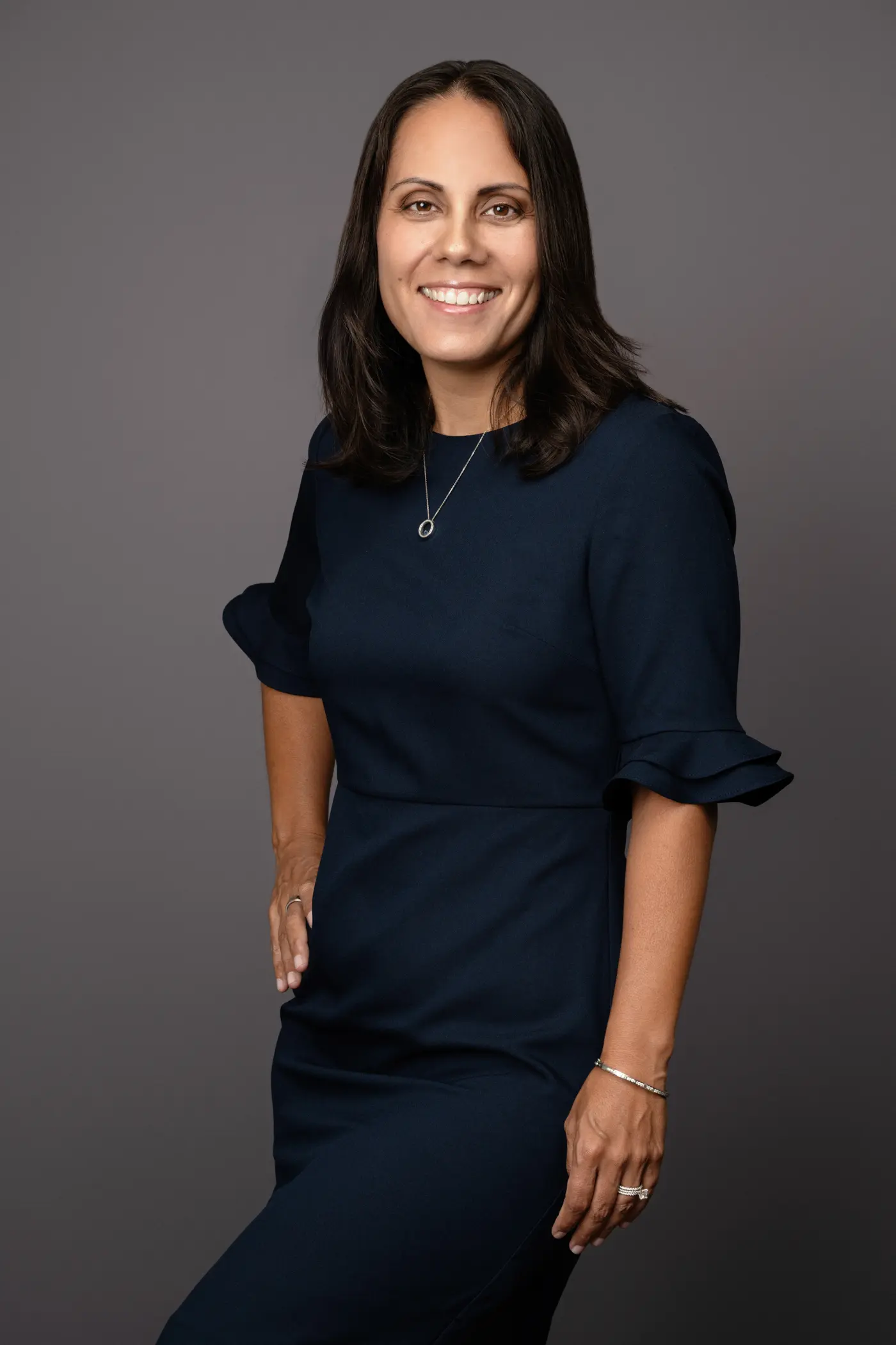Smiling woman in a navy blue dress with rolled sleeves against a gray background.